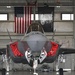 A front-facing view of an F-35 Lightning II stealth fighter jet parked inside a military hangar. The aircraft has its weapon bay doors open, revealing red safety covers and 'Remove Before Flight' tags. An American flag and a black 'WEAPONS' banner are prominently displayed on the hangar wall behind the aircraft.