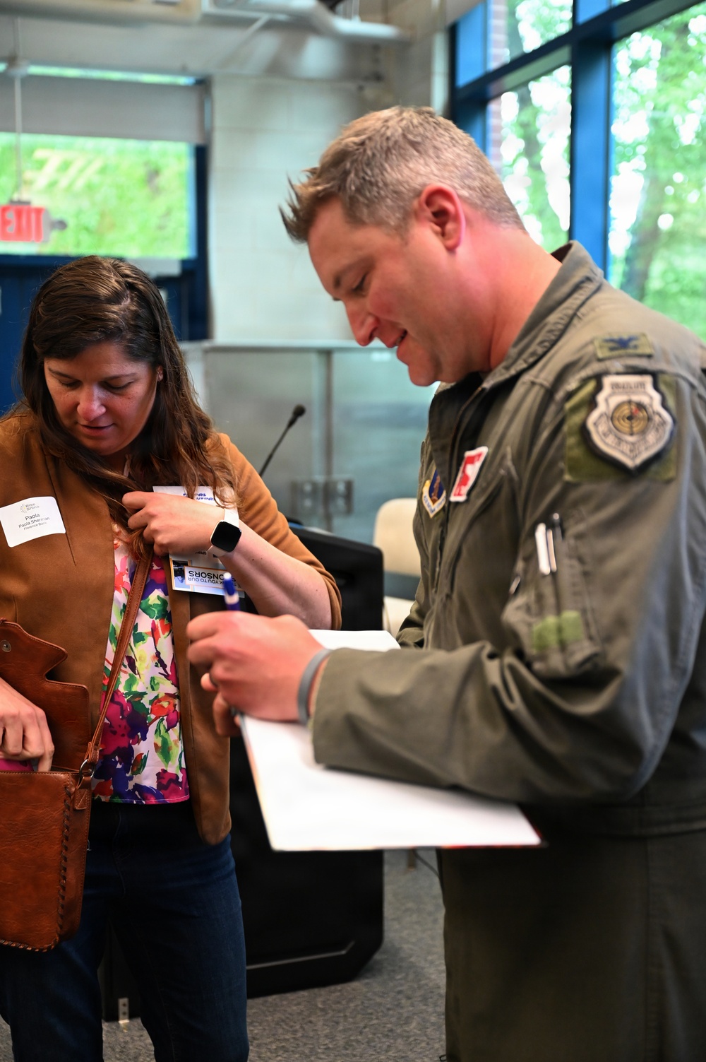 A military officer in an olive green flight suit is interacting with a civilian woman at what appears to be an indoor event or meeting. The officer is holding a clipboard or document and appears to be writing or reviewing information, while the woman, wearing a name badge, looks on. The setting appears to be a conference room or event space.