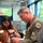 104th Fighter Wing hosts Springfield Chamber of Commerce Breakfast A military officer in an olive green flight suit is interacting with a civilian woman at what appears to be an indoor event or meeting. The officer is holding a clipboard or document and appears to be writing or reviewing information, while the woman, wearing a name badge, looks on. The setting appears to be a conference room or event space.