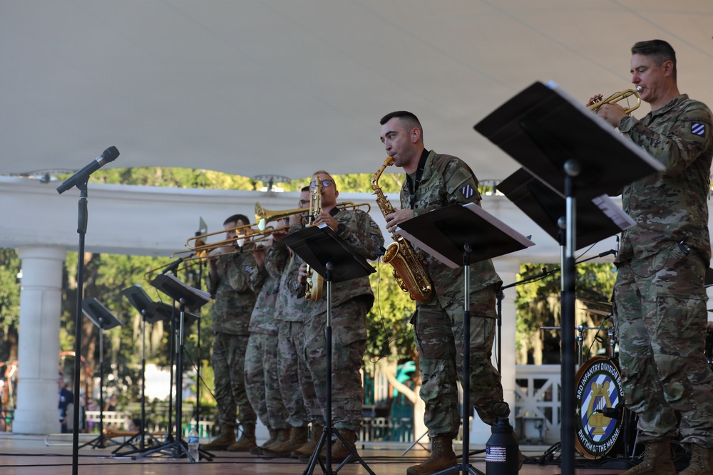 Military musicians in U.S. Army camouflage uniforms perform on an outdoor stage under a white tent canopy. The ensemble appears to be the 3rd Infantry Division Band, as indicated by the bass drum head visible in the background. Musicians are playing brass and woodwind instruments with sheet music on stands.