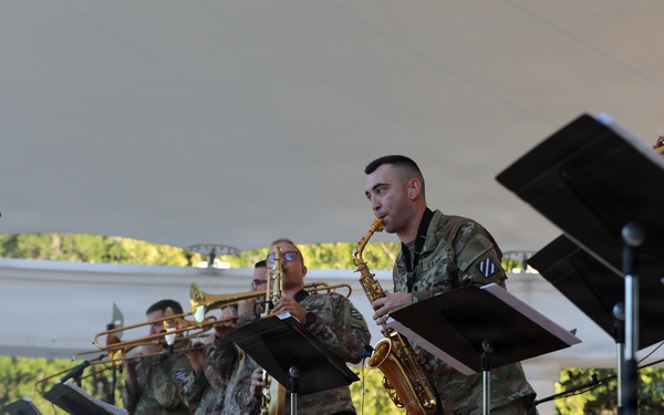 Military musicians in U.S. Army camouflage uniforms perform on an outdoor stage under a white tent canopy. The ensemble appears to be the 3rd Infantry Division Band, as indicated by the bass drum head visible in the background. Musicians are playing brass and woodwind instruments with sheet music on stands.