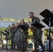 Military musicians in U.S. Army camouflage uniforms perform on an outdoor stage under a white tent canopy. The ensemble appears to be the 3rd Infantry Division Band, as indicated by the bass drum head visible in the background. Musicians are playing brass and woodwind instruments with sheet music on stands.