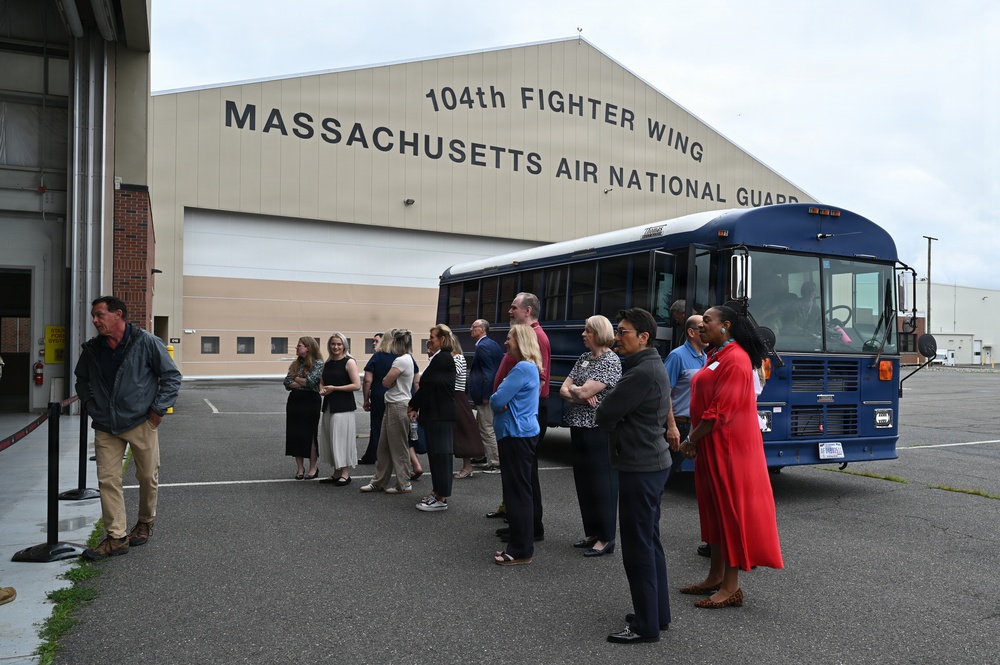 A group of civilians is gathered in a parking area outside the 104th Fighter Wing, Massachusetts Air National Guard hangar facility. They appear to be on an organized visit or tour, standing in a loose line alongside a blue bus. The setting suggests an official community outreach, employer support, or civic leader tour event.