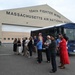 A group of civilians is gathered in a parking area outside the 104th Fighter Wing, Massachusetts Air National Guard hangar facility. They appear to be on an organized visit or tour, standing in a loose line alongside a blue bus. The setting suggests an official community outreach, employer support, or civic leader tour event.