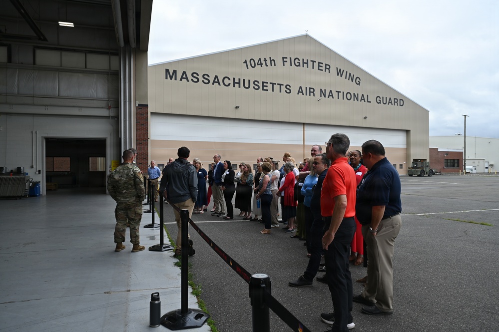 A group of civilian visitors is gathered outside a large military hangar belonging to the 104th Fighter Wing, Massachusetts Air National Guard. A uniformed military personnel member appears to be briefing or addressing the group, which is cordoned off by stanchion barriers. The event appears to be an organized tour or community outreach visit to the air base.