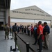 A group of civilian visitors is gathered outside a large military hangar belonging to the 104th Fighter Wing, Massachusetts Air National Guard. A uniformed military personnel member appears to be briefing or addressing the group, which is cordoned off by stanchion barriers. The event appears to be an organized tour or community outreach visit to the air base.