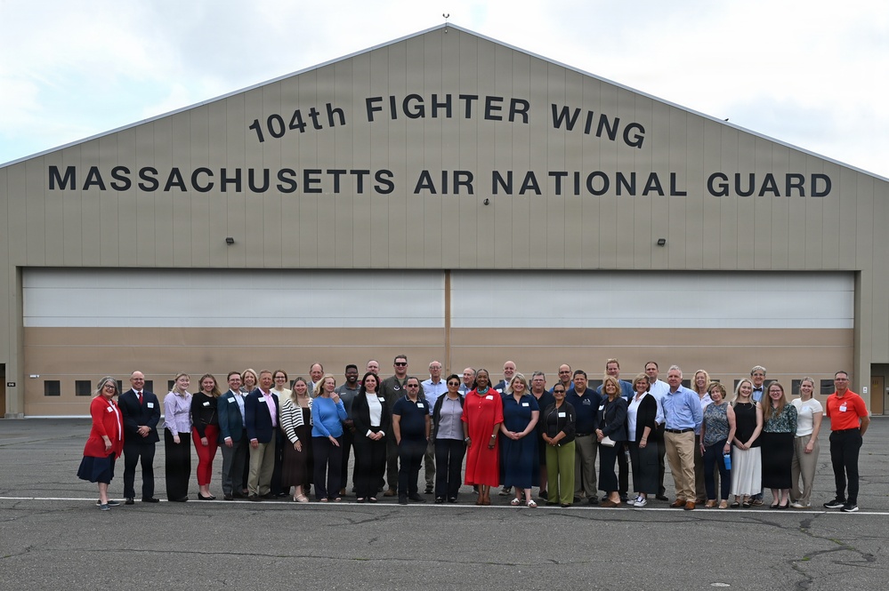 A large group of approximately 35-40 civilians and possibly some military personnel in civilian attire pose for a group photograph in front of a large hangar belonging to the 104th Fighter Wing, Massachusetts Air National Guard. The group appears to be participating in a community outreach, employer support, or civic leader visit event at the military installation. The setting is an outdoor tarmac area in front of the hangar's large bay doors.