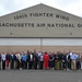 A large group of approximately 35-40 civilians and possibly some military personnel in civilian attire pose for a group photograph in front of a large hangar belonging to the 104th Fighter Wing, Massachusetts Air National Guard. The group appears to be participating in a community outreach, employer support, or civic leader visit event at the military installation. The setting is an outdoor tarmac area in front of the hangar's large bay doors.