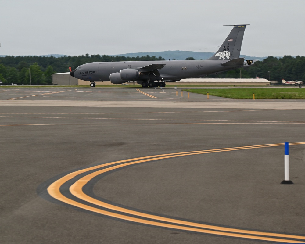 A U.S. Air Force KC-135 Stratotanker aircraft with tail code 'AK' and serial number 0332 is taxiing on an airfield tarmac. The aircraft bears Alaska Air National Guard markings, identifiable by the polar bear emblem on the tail fin. The setting appears to be a regional or military airfield surrounded by forested hills under overcast skies.