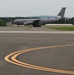 A U.S. Air Force KC-135 Stratotanker aircraft with tail code 'AK' and serial number 0332 is taxiing on an airfield tarmac. The aircraft bears Alaska Air National Guard markings, identifiable by the polar bear emblem on the tail fin. The setting appears to be a regional or military airfield surrounded by forested hills under overcast skies.