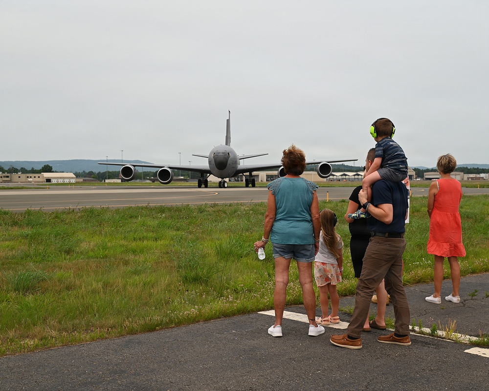 A group of civilians, appearing to be a family, stands at the edge of a military airfield watching a large gray military aircraft taxi on the runway. The scene suggests a deployment or homecoming event, with family members of varying ages gathered to observe the aircraft. The setting is a military air base with hangars and infrastructure visible in the background.