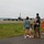 104th Fighter Wing Celebrates Flight Surgeon's Fifteen Years of Service with Fini Flight A group of civilians, appearing to be a family, stands at the edge of a military airfield watching a large gray military aircraft taxi on the runway. The scene suggests a deployment or homecoming event, with family members of varying ages gathered to observe the aircraft. The setting is a military air base with hangars and infrastructure visible in the background.