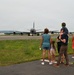 A group of civilians, appearing to be a family, stands at the edge of a military airfield watching a large gray military aircraft taxi on the runway. The scene suggests a deployment or homecoming event, with family members of varying ages gathered to observe the aircraft. The setting is a military air base with hangars and infrastructure visible in the background.