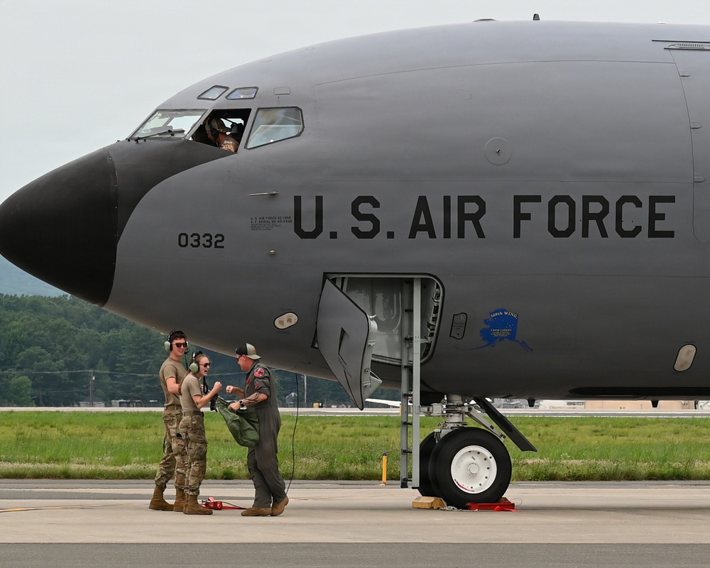 A U.S. Air Force military aircraft, identified by the serial number 0332, is parked on a tarmac. Three ground crew members in military uniforms are standing near the nose landing gear, appearing to communicate with each other. A pilot or crew member is visible in the cockpit window above.