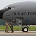 A U.S. Air Force military aircraft, identified by the serial number 0332, is parked on a tarmac. Three ground crew members in military uniforms are standing near the nose landing gear, appearing to communicate with each other. A pilot or crew member is visible in the cockpit window above.