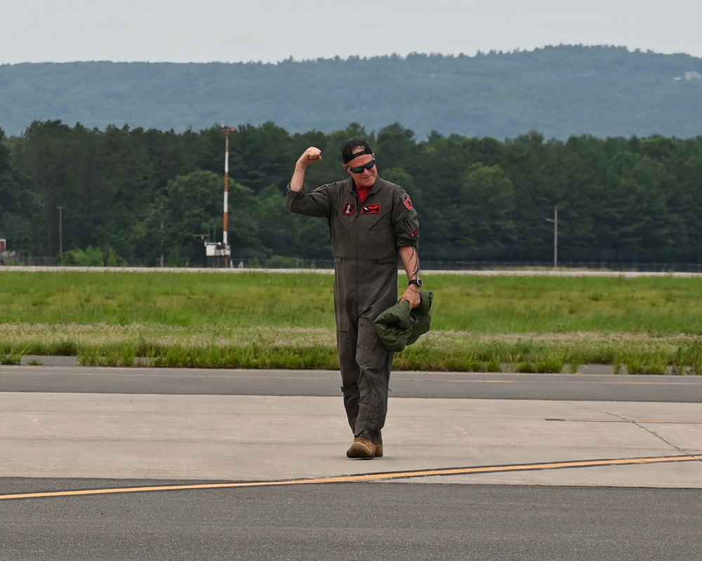A military aviator in an olive drab flight suit walks confidently across an airfield tarmac while raising his fist in a celebratory gesture. He is carrying what appears to be a flight helmet in his left hand and wearing sunglasses and a backwards cap. The background shows a runway environment with green treeline and hills in the distance.