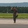 104th Fighter Wing Celebrates Flight Surgeon's Fifteen Years of Service with Fini Flight A military aviator in an olive drab flight suit walks confidently across an airfield tarmac while raising his fist in a celebratory gesture. He is carrying what appears to be a flight helmet in his left hand and wearing sunglasses and a backwards cap. The background shows a runway environment with green treeline and hills in the distance.