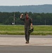 A military aviator in an olive drab flight suit walks confidently across an airfield tarmac while raising his fist in a celebratory gesture. He is carrying what appears to be a flight helmet in his left hand and wearing sunglasses and a backwards cap. The background shows a runway environment with green treeline and hills in the distance.