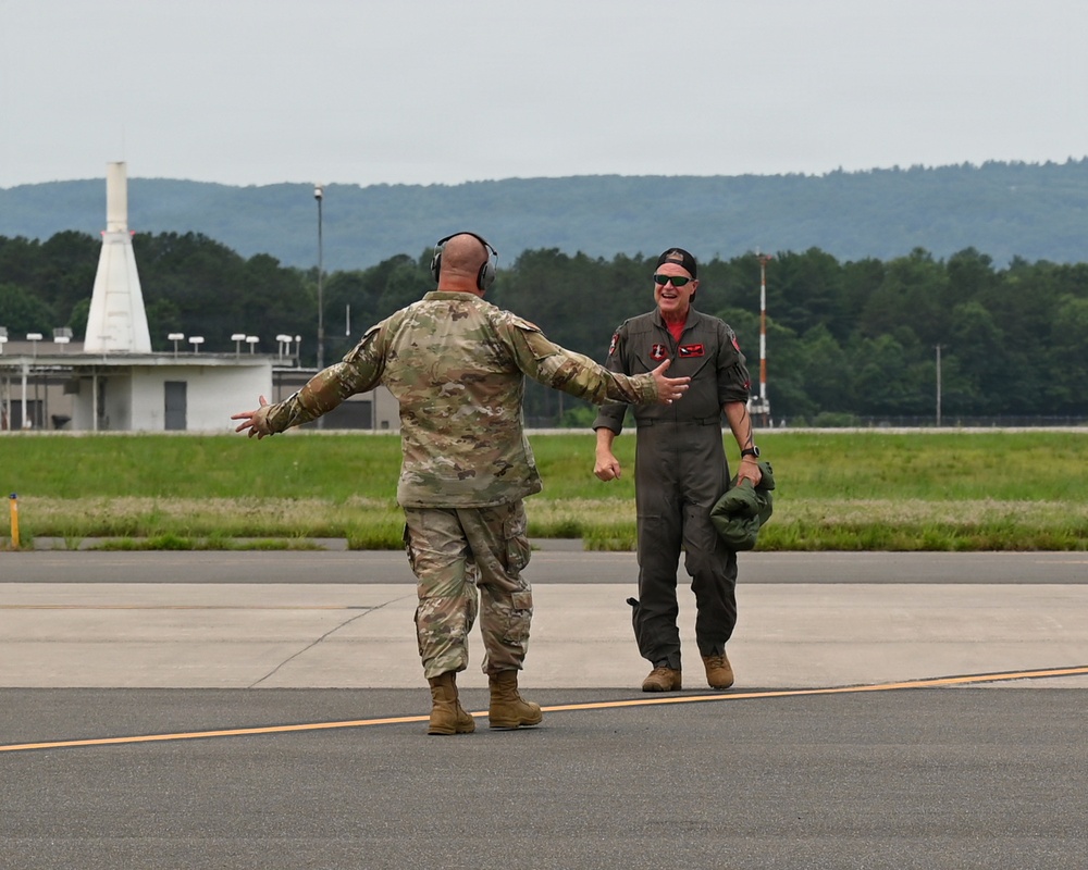 Two military personnel are greeting each other on an airfield tarmac. One individual is wearing camouflage combat uniform with hearing protection, while the other is dressed in an olive flight suit and carrying a helmet. Both appear to be in a welcoming or celebratory interaction, possibly following a flight mission.