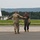 104th Fighter Wing Celebrates Flight Surgeon's Fifteen Years of Service with Fini Flight Two military personnel are greeting each other on an airfield tarmac. One individual is wearing camouflage combat uniform with hearing protection, while the other is dressed in an olive flight suit and carrying a helmet. Both appear to be in a welcoming or celebratory interaction, possibly following a flight mission.