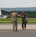 Two military personnel are greeting each other on an airfield tarmac. One individual is wearing camouflage combat uniform with hearing protection, while the other is dressed in an olive flight suit and carrying a helmet. Both appear to be in a welcoming or celebratory interaction, possibly following a flight mission.