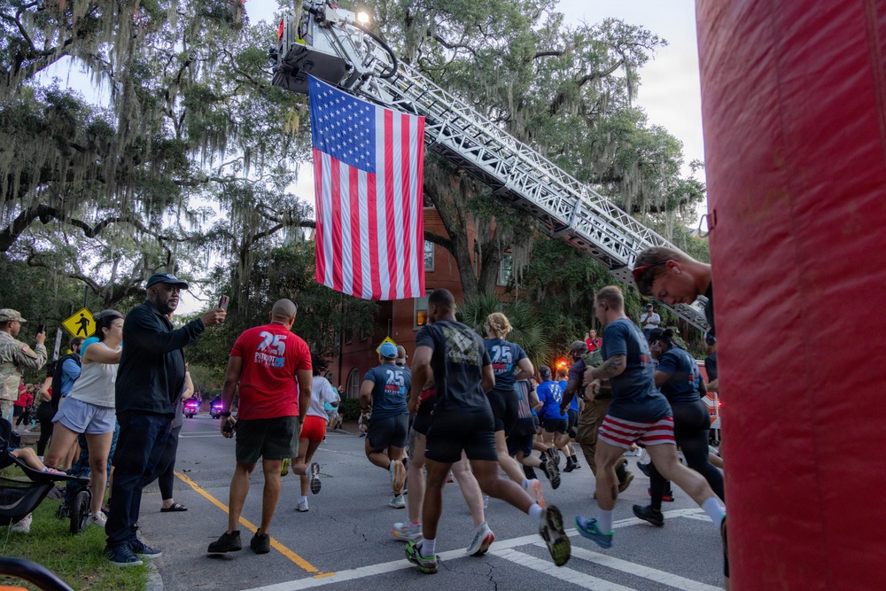 A group of runners participates in what appears to be a patriotic road race or fun run, passing beneath a large American flag suspended from an extended fire truck ladder. The event takes place on a tree-lined street with Spanish moss-draped oak trees, suggesting a Southern U.S. location. Spectators line the sidewalk cheering on participants, many of whom wear matching numbered race shirts.