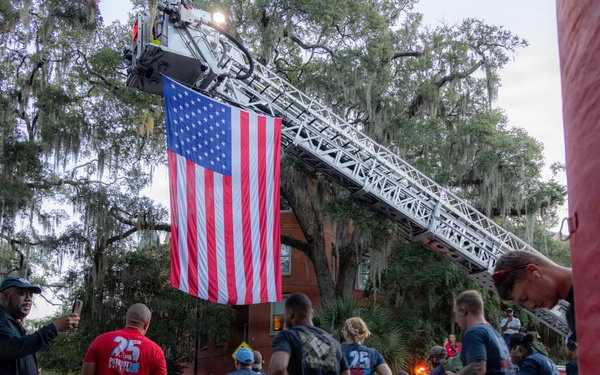 A group of runners participates in what appears to be a patriotic road race or fun run, passing beneath a large American flag suspended from an extended fire truck ladder. The event takes place on a tree-lined street with Spanish moss-draped oak trees, suggesting a Southern U.S. location. Spectators line the sidewalk cheering on participants, many of whom wear matching numbered race shirts.