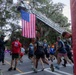 A group of runners participates in what appears to be a patriotic road race or fun run, passing beneath a large American flag suspended from an extended fire truck ladder. The event takes place on a tree-lined street with Spanish moss-draped oak trees, suggesting a Southern U.S. location. Spectators line the sidewalk cheering on participants, many of whom wear matching numbered race shirts.