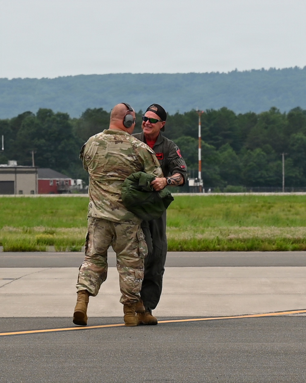 Two individuals are interacting on a military airfield tarmac. One person is wearing a military camouflage (OCP) uniform with hearing protection, while the other is wearing an olive drab flight suit with a red shirt underneath, sunglasses, and a backwards cap. They appear to be engaged in a friendly greeting or handshake on the flight line.