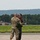 104th Fighter Wing Celebrates Flight Surgeon's Fifteen Years of Service with Fini Flight Two individuals are interacting on a military airfield tarmac. One person is wearing a military camouflage (OCP) uniform with hearing protection, while the other is wearing an olive drab flight suit with a red shirt underneath, sunglasses, and a backwards cap. They appear to be engaged in a friendly greeting or handshake on the flight line.
