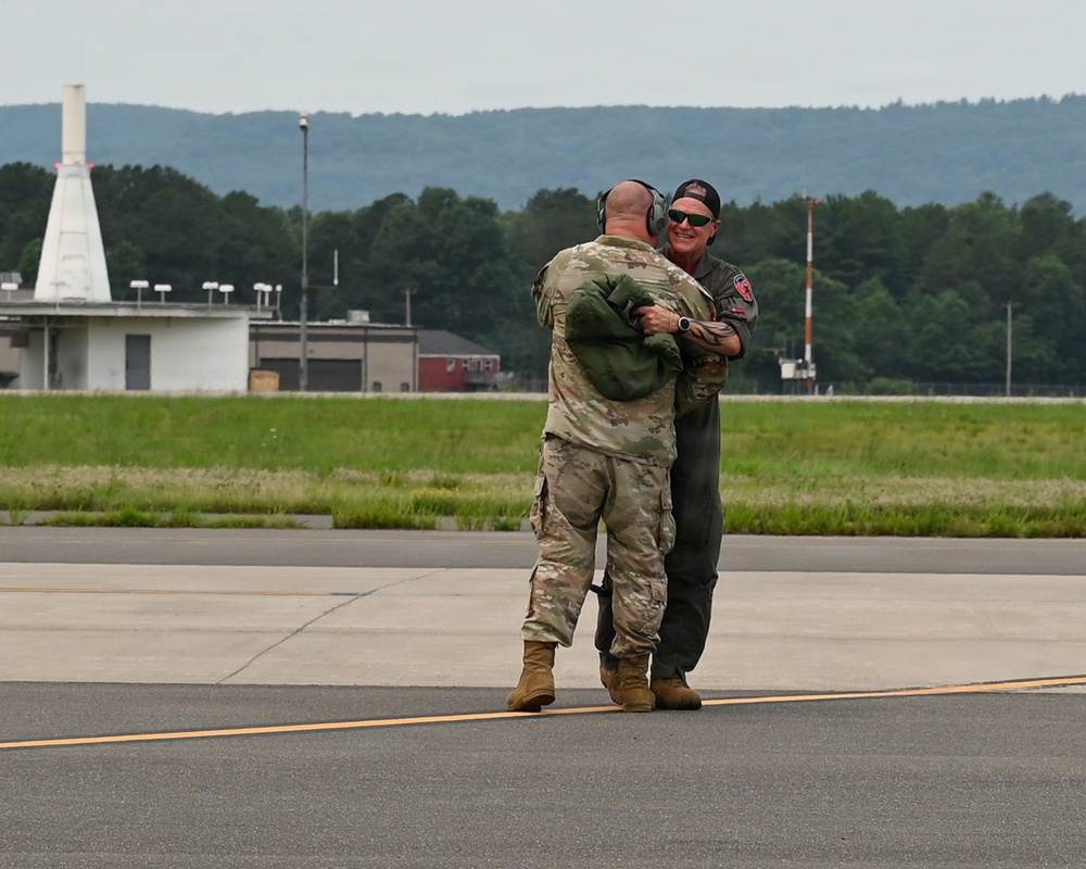 Two military personnel are embracing on an airfield tarmac, suggesting a celebratory or farewell moment. One individual is wearing camouflage (OCP/MultiCam) uniform with hearing protection, while the other wears a dark flight suit with a baseball cap and sunglasses. The setting appears to be a military or joint-use airfield with hangars, a control tower marker, and wooded hills visible in the background.
