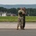 104th Fighter Wing Celebrates Flight Surgeon's Fifteen Years of Service with Fini Flight Two military personnel are embracing on an airfield tarmac, suggesting a celebratory or farewell moment. One individual is wearing camouflage (OCP/MultiCam) uniform with hearing protection, while the other wears a dark flight suit with a baseball cap and sunglasses. The setting appears to be a military or joint-use airfield with hangars, a control tower marker, and wooded hills visible in the background.