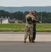 Two military personnel are embracing on an airfield tarmac, suggesting a celebratory or farewell moment. One individual is wearing camouflage (OCP/MultiCam) uniform with hearing protection, while the other wears a dark flight suit with a baseball cap and sunglasses. The setting appears to be a military or joint-use airfield with hangars, a control tower marker, and wooded hills visible in the background.