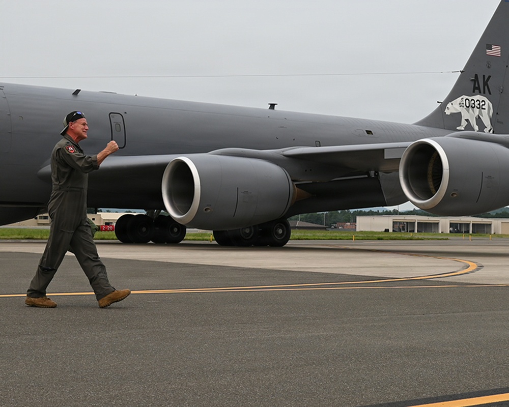 A military aviator in olive green flight suit walks across a tarmac in front of a large gray military tanker aircraft. The aircraft displays 'AK' tail codes, a polar bear emblem, and the serial number 0332, identifying it as an Alaska-based KC-135 Stratotanker. The pilot appears to be in good spirits, gesturing with a raised fist.