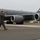 104th Fighter Wing Celebrates Flight Surgeon's Fifteen Years of Service with Fini Flight A military aviator in olive green flight suit walks across a tarmac in front of a large gray military tanker aircraft. The aircraft displays 'AK' tail codes, a polar bear emblem, and the serial number 0332, identifying it as an Alaska-based KC-135 Stratotanker. The pilot appears to be in good spirits, gesturing with a raised fist.