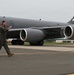 A military aviator in olive green flight suit walks across a tarmac in front of a large gray military tanker aircraft. The aircraft displays 'AK' tail codes, a polar bear emblem, and the serial number 0332, identifying it as an Alaska-based KC-135 Stratotanker. The pilot appears to be in good spirits, gesturing with a raised fist.