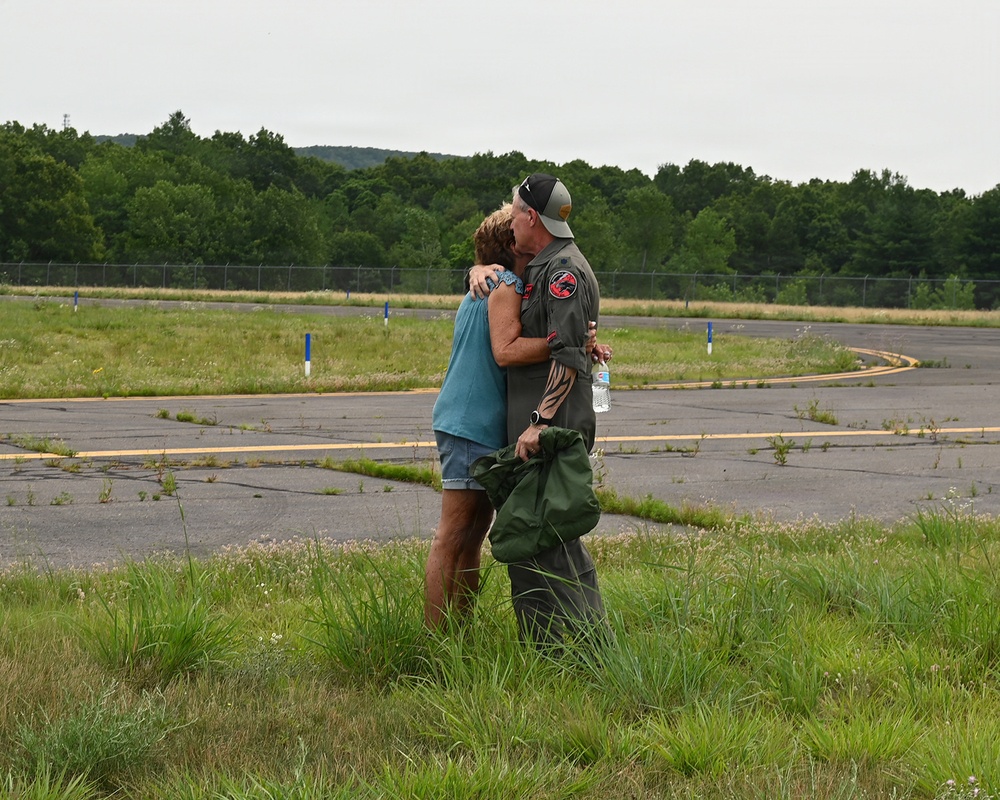 Two people are embracing and kissing on what appears to be an airfield or tarmac area. One individual is wearing a military or aviation flight suit with patches, a cap, and is holding a green bag and water bottle. The setting appears to be a reunion or homecoming moment at an airfield surrounded by trees and overgrown grass.