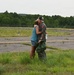 Two people are embracing and kissing on what appears to be an airfield or tarmac area. One individual is wearing a military or aviation flight suit with patches, a cap, and is holding a green bag and water bottle. The setting appears to be a reunion or homecoming moment at an airfield surrounded by trees and overgrown grass.