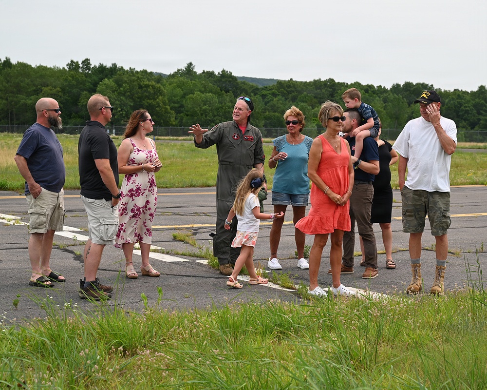 A group of approximately 11 people, including adults and two children, are gathered on an airfield tarmac. A person wearing an olive-green flight suit with patches is speaking and gesturing to the group, suggesting a briefing or tour. The setting appears to be a small regional or military airfield surrounded by trees and open grass fields.