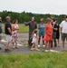 A group of approximately 11 people, including adults and two children, are gathered on an airfield tarmac. A person wearing an olive-green flight suit with patches is speaking and gesturing to the group, suggesting a briefing or tour. The setting appears to be a small regional or military airfield surrounded by trees and open grass fields.