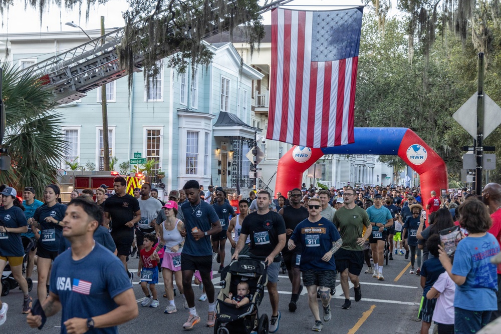 A large outdoor road race or fun run is underway on a tree-lined urban street, with hundreds of participants running beneath a large American flag suspended from a fire truck ladder. The event appears to be organized by MWR (Morale, Welfare, and Recreation), suggesting a military-affiliated community event. Participants wear race bibs numbered '911', indicating a possible first responder or patriotic-themed race.