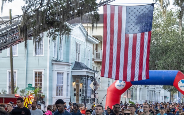 A large outdoor road race or fun run is underway on a tree-lined urban street, with hundreds of participants running beneath a large American flag suspended from a fire truck ladder. The event appears to be organized by MWR (Morale, Welfare, and Recreation), suggesting a military-affiliated community event. Participants wear race bibs numbered '911', indicating a possible first responder or patriotic-themed race.