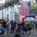 A large outdoor road race or fun run is underway on a tree-lined urban street, with hundreds of participants running beneath a large American flag suspended from a fire truck ladder. The event appears to be organized by MWR (Morale, Welfare, and Recreation), suggesting a military-affiliated community event. Participants wear race bibs numbered '911', indicating a possible first responder or patriotic-themed race.
