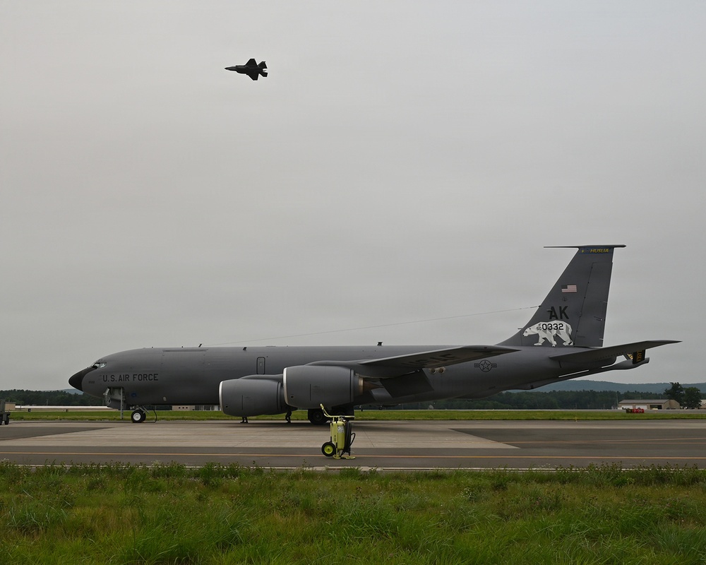A U.S. Air Force KC-135 Stratotanker aerial refueling aircraft is parked on a taxiway or ramp at a military airfield. The aircraft bears 'AK' tail codes and serial number '0332', indicating Alaska Air National Guard assignment, with a polar bear emblem visible on the tail. A fighter jet, appearing to be an F-35, is visible flying overhead in the overcast sky.
