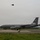 104th Fighter Wing Celebrates Flight Surgeon's Fifteen Years of Service with Fini Flight A U.S. Air Force KC-135 Stratotanker aerial refueling aircraft is parked on a taxiway or ramp at a military airfield. The aircraft bears 'AK' tail codes and serial number '0332', indicating Alaska Air National Guard assignment, with a polar bear emblem visible on the tail. A fighter jet, appearing to be an F-35, is visible flying overhead in the overcast sky.
