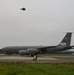 A U.S. Air Force KC-135 Stratotanker aerial refueling aircraft is parked on a taxiway or ramp at a military airfield. The aircraft bears 'AK' tail codes and serial number '0332', indicating Alaska Air National Guard assignment, with a polar bear emblem visible on the tail. A fighter jet, appearing to be an F-35, is visible flying overhead in the overcast sky.