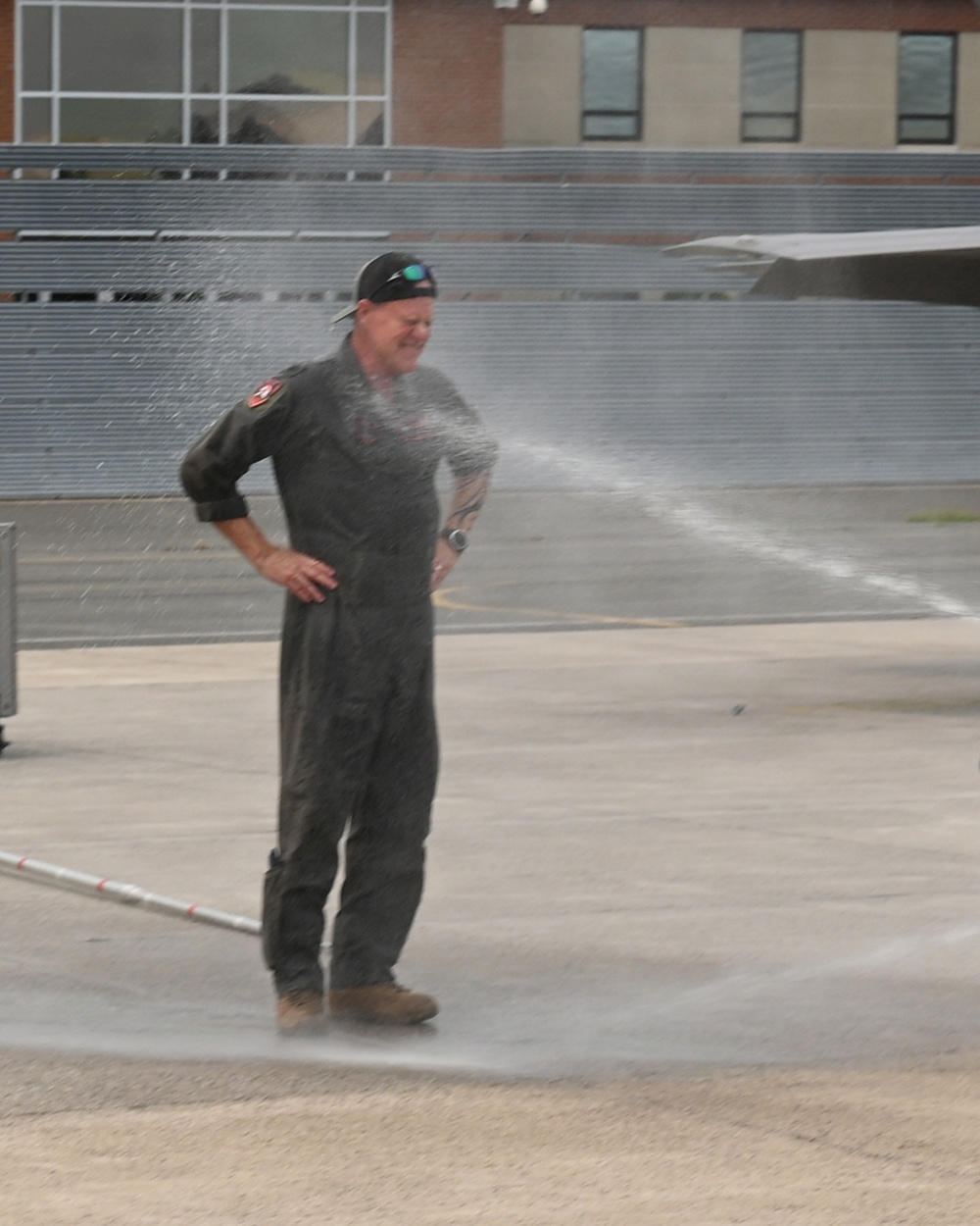 A person wearing a dark flight suit and baseball cap is being sprayed with water on what appears to be a military airfield tarmac. The individual is smiling and standing with hands on hips, suggesting this is a celebratory tradition. This appears consistent with a military aviation 'fini flight' or retirement water salute ceremony.