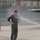 104th Fighter Wing Celebrates Flight Surgeon's Fifteen Years of Service with Fini Flight A person wearing a dark flight suit and baseball cap is being sprayed with water on what appears to be a military airfield tarmac. The individual is smiling and standing with hands on hips, suggesting this is a celebratory tradition. This appears consistent with a military aviation 'fini flight' or retirement water salute ceremony.