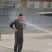 A person wearing a dark flight suit and baseball cap is being sprayed with water on what appears to be a military airfield tarmac. The individual is smiling and standing with hands on hips, suggesting this is a celebratory tradition. This appears consistent with a military aviation 'fini flight' or retirement water salute ceremony.