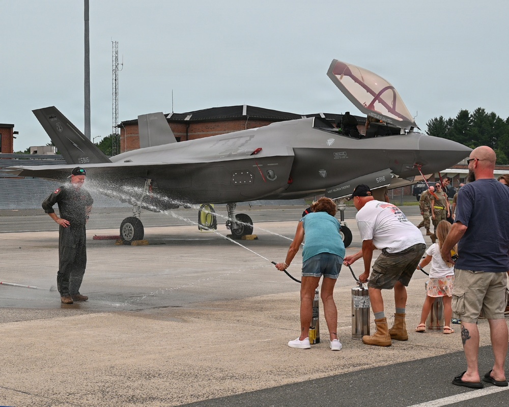 A military pilot in a flight suit is being sprayed with water by family members on an airfield tarmac in front of an F-35 Lightning II fighter jet bearing tail number 5816. This appears to be a traditional 'fini flight' or final flight celebration, a military aviation tradition where a pilot completing their last flight is doused with water. A small group of family members, including children, and military personnel in the background are participating in the ceremony.